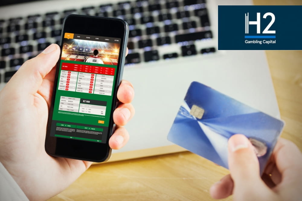 Il logo di H2 Gambling Capital, uno smartphone con la schermata di un sito di betting online, una carta di credito e la tastiera di un laptop su una scrivania